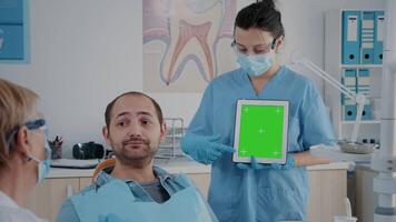 Assistant vertically holding digital tablet with green screen at appointment with patient in cabinet. Woman pointing at mock up template with blank copy space and isolated chroma key. video