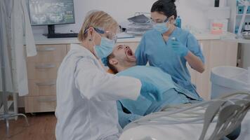 Mannschaft von Spezialisten tun bohren Verfahren auf geduldig mit Zahnschmerzen, mit Dental Instrumente im Oral Pflege Büro. Medizin und Assistent tun Karies Inspektion, Behandlung Mann im Schmerz. video