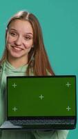 Vertical Smiling woman presenting laptop with green screen display, isolated over blue studio background. Cheerful person creating promotion with chroma key notebook device, camera A video