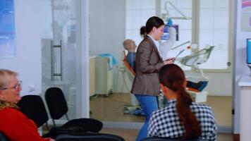 Young lady asking informations filling in stomatological form while patients talking sitting on chair in waiting area. People speaking in crowded professional orthodontist reception office. video