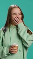 Vertical Young girl with backpack yawning, feeling tired, drinking coffee to wake up. Sleepy woman using caffeinated beverage to gain energy, lacking sleep, studio background, camera A video