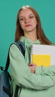 verticale portrait de joyeux adolescent avec sac à dos en portant école carnet et Remarques, isolé plus de studio Contexte. gai élève avec école Provisions dans bras, en train de préparer à aller à collège, caméra une video