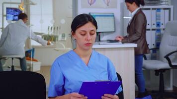 Portrait of smiling nurse in dental office while doctor is working with patient in background. Stomatologist assistant looking on webcam sitting on chair in stomatological clinic. video