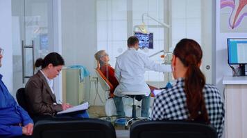 Patients sitting on chairs in waiting room of stomatological clinic filling in stomatological forms while doctor working in background. Concept of crowded professional orthodontist reception office. video