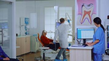 Dentist inviting senior man in consultation dental room while nurse giving to patient form to fill indicating to sit on chair in waiting area. Slow motion shot crowded professional orthodontist office video