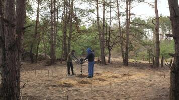 On an autumn morning, builders drill the soil in a forest clearing to install a concrete foundation. The ground is marked with several small stakes, indicating a methodical approach to their work. video