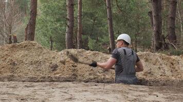 A worker wearing a white hard hat is actively digging a narrow trench in a forested area. The surrounding terrain is uneven with visible tree stumps and soil heaps. video