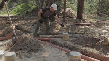 un' lavoratore, rivestito nel opera abbigliamento e attrezzata con sicurezza Ingranaggio, è nel il processi di posa grande metropolitana tubi tra il parzialmente sepolto calcestruzzo fondamenta nel un' foreste costruzione luogo. video