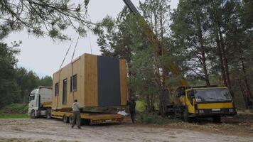 A crane is lifting a prefabricated house from a trailer in a forest clearing during an early morning. Workers are present to guide the operation. video