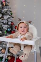 chubby baby in a jumpsuit with deer horns sits in a white high chair for feeding near Christmas tree photo