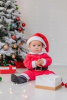 baby in a Santa costume is sitting next to the Christmas tree and playing with gift boxes. lifestyle photo