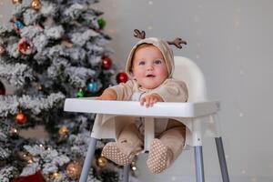 gordito bebé en un mono con ciervo cuernos se sienta en un blanco alto silla para alimentación cerca Navidad árbol foto