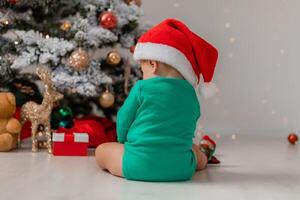 bebé en gnomo disfraz es sentado en el piso siguiente a el Navidad árbol con su espalda a el cámara foto