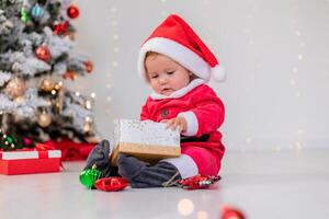 baby in a Santa costume is sitting next to the Christmas tree and playing with gift boxes. lifestyle photo