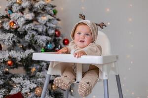 chubby baby in a jumpsuit with deer horns sits in a white high chair for feeding near Christmas tree photo