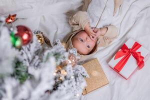 cute baby in a bodysuit is lying on the bed next to a Christmas tree and gift boxes. top view photo