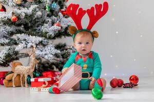 baby in a green bodysuit with red deer horns on his head is playing next to the Christmas tree photo