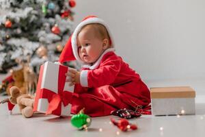 baby in a Santa costume is sitting next to the Christmas tree and playing with gift boxes. lifestyle photo