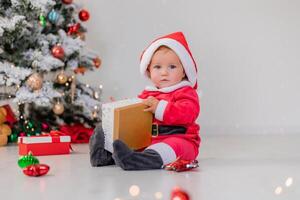 baby in a Santa costume is sitting next to the Christmas tree and playing with gift boxes. lifestyle photo