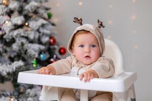 portrait of chubby baby in a jumpsuit with deer horns sits in a white high chair near Christmas tree photo