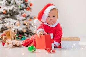 baby in a Santa costume is sitting next to the Christmas tree and playing with gift boxes. lifestyle photo