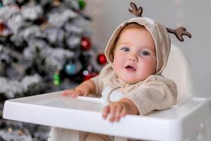 portrait of chubby baby in a jumpsuit with deer horns sits in a white high chair near Christmas tree photo