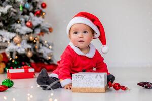 baby in a Santa costume is sitting next to the Christmas tree and playing with gift boxes. lifestyle photo