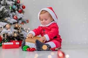 baby in a Santa costume is sitting next to the Christmas tree and playing with gift boxes. lifestyle photo