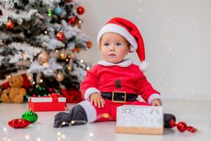 baby in a Santa costume is sitting next to the Christmas tree and playing with gift boxes. lifestyle photo