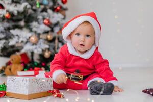 baby in a Santa costume is sitting next to the Christmas tree and playing with gift boxes. lifestyle photo