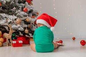 bebé en gnomo disfraz es sentado en el piso siguiente a el Navidad árbol con su espalda a el cámara foto