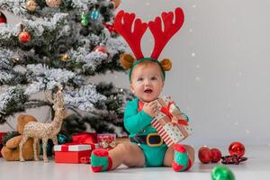 baby in a green bodysuit with red deer horns on his head is playing next to the Christmas tree photo