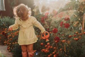 pequeño niña en un amarillo vestir y caucho botas es riego flores en el otoño jardín foto