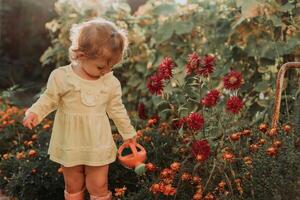 pequeño niña en un amarillo vestir y caucho botas es riego flores en el otoño jardín foto