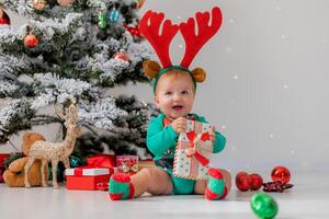 baby in a green bodysuit with red deer horns on his head is playing next to the Christmas tree photo