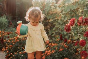 pequeño niña en un amarillo vestir y caucho botas es riego flores en el otoño jardín foto