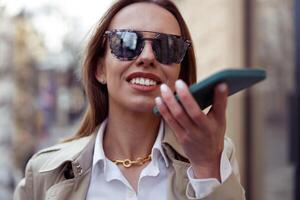 Happy businesswoman in sunglasses is recording audio message on phone on city street background photo