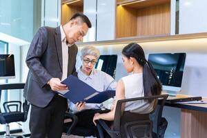Asian senior business woman giving advice to the new manager and looking at the information regarding sale record while working in modern style office photo