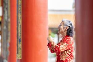 asiático mayor mujer en rojo cheongsam qipao vestir es haciendo un deseo a ancestral Dios dentro chino budista templo durante lunar nuevo año para mejor deseo bendición y bueno suerte foto