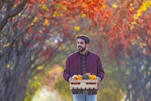 Happy caucasian farmer carrying organics homegrown produce harvest with apple, squash and pumpkin while walking along the country road with fall color from maple and beech tree during autumn season photo
