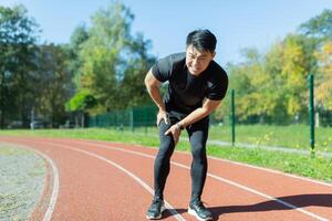 joven asiático corredor atleta con músculo dolor. hombre masajear extensión, trauma lesión mientras trotar a el estadio al aire libre. aptitud masculino esguince grave dolor tramo jalar. pierna músculo calambre becerro deporte foto