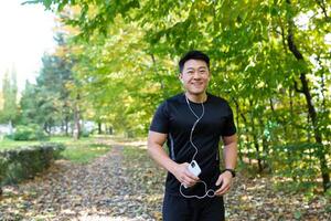 Portrait of happy sportsman in park, Asian man running and listening to music in headphones, using online application for listening to radio and podcasts, looking at camera and smiling photo