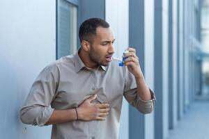 A man outside an office building has a severe asthma attack, a businessman is having difficulty breathing, a worker in a shirt is using an inhaler to make breathing easier, a hispanic man in a casual shirt. photo