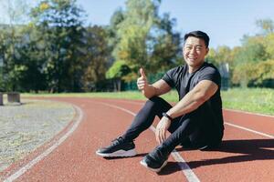 retrato de un asiático deportista sentado en un estadio rueda de andar en un público parque en un verano soleado día, el hombre es mirando a el cámara y sonriente dando pulgares arriba, corredor en ropa de deporte afuera. foto