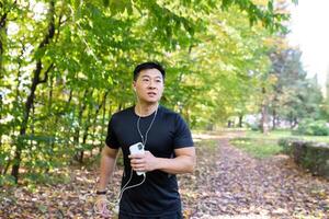 asiático deportista Mañana correr, hombre con auriculares escuchando a música y audiolibros con podcasts, deportista en otoño parque corriendo en soleado día. foto