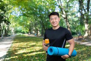 Fitness trainer looking at camera and smiling, asian sports instructor with sports mat and bottle of water, in autumn park on sunny day near trees. photo