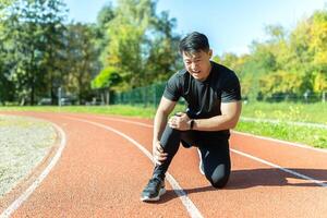 Asian man after fitness workout and jogging sits on the ground and suffers from leg pain, massages leg muscles photo