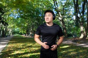 asiático deportista Mañana correr, hombre con auriculares escuchando a música y audiolibros con podcasts, deportista en otoño parque corriendo en soleado día. foto