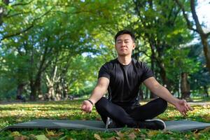 Portrait of Asian man in autumn park meditating sitting in lotus position on fitness mat, sportsman resting after training doing breathing exercises, photo