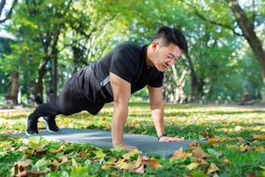 asiático atleta haciendo Lagartijas en parque y haciendo tablón en Deportes estera, hombre contento con rutina de ejercicio en soleado día. foto